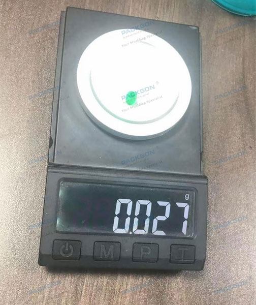 Packson micro-injection molding excellence, showcasing a tiny green component on a digital scale weighing only 0.027 grams to demonstrate extreme manufacturing precision and tight tolerances.